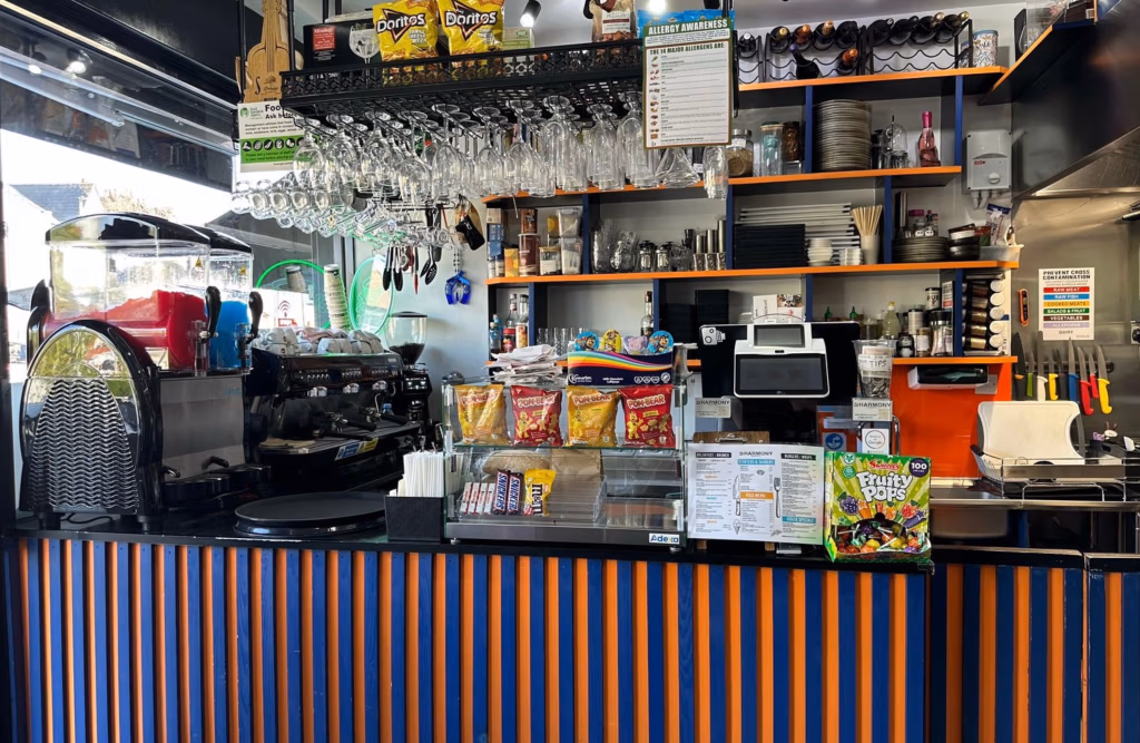 Refurbished café service counter with espresso machine, hanging wine glasses, orange and blue panelled front, shelves stocked with snacks and café supplies