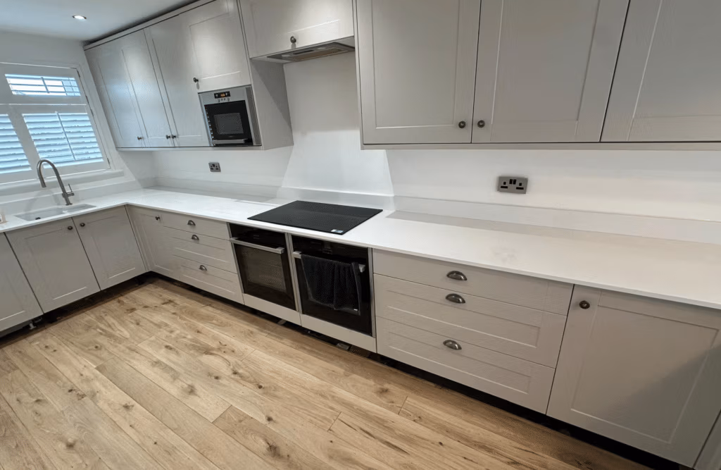 Renovated kitchen with light grey shaker cabinets, white quartz worktops, black induction hob, stainless steel oven, and oak-effect flooring