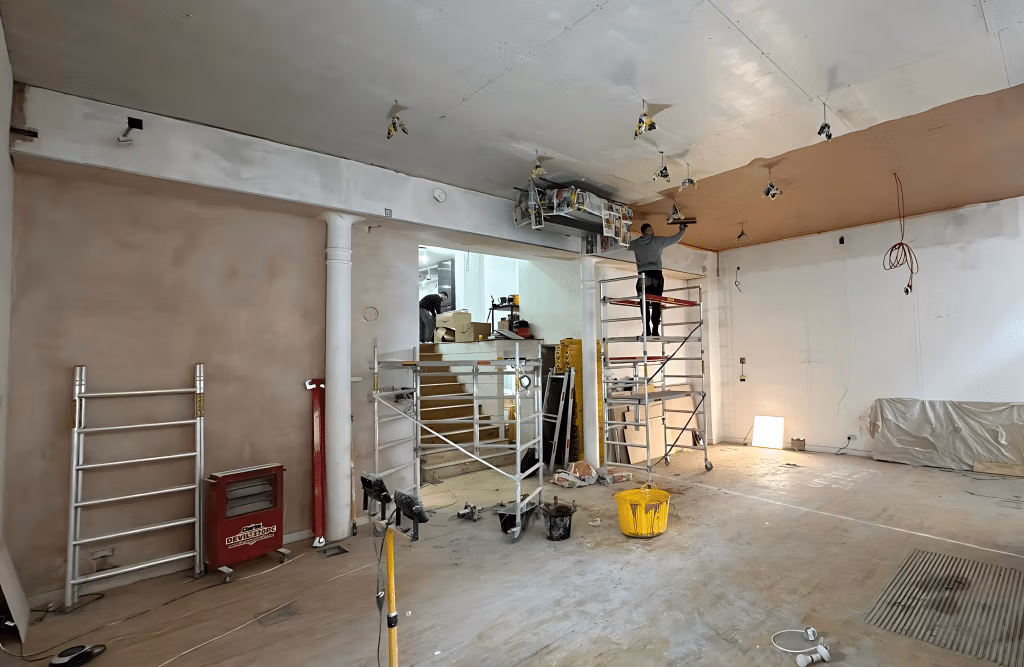 Large commercial space mid-renovation with workers on scaffolding installing ceiling fixtures, partially plastered walls, and construction equipment throughout